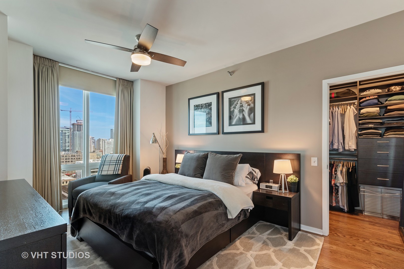 Staged condo bedroom with gray bedding, a walk-in closet, and city views through tall windows.