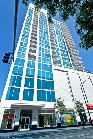 Modern high-rise residential building with blue glass windows and white balconies, viewed from the street.