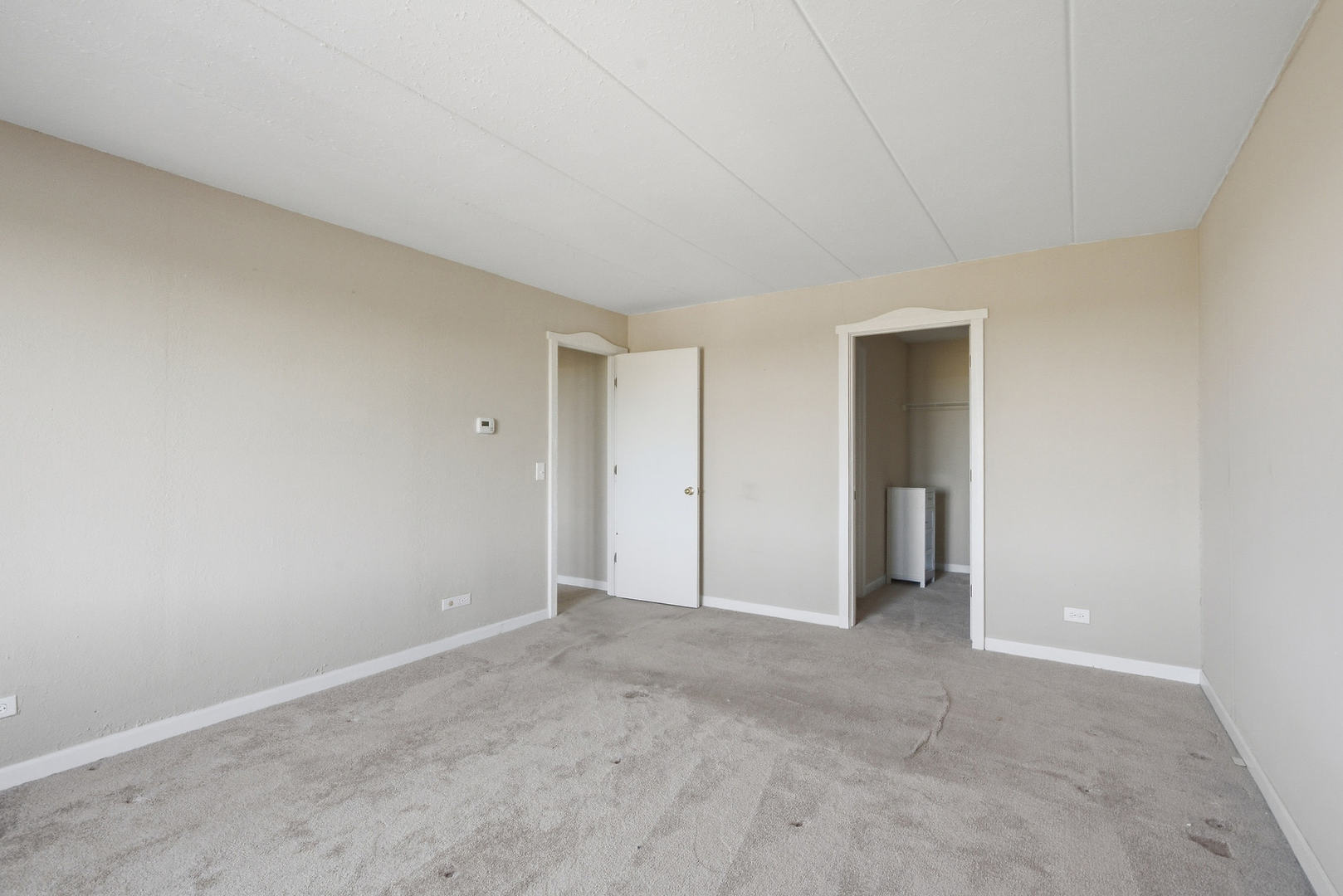 Empty carpeted room with beige walls and two doorways, including an open closet.
