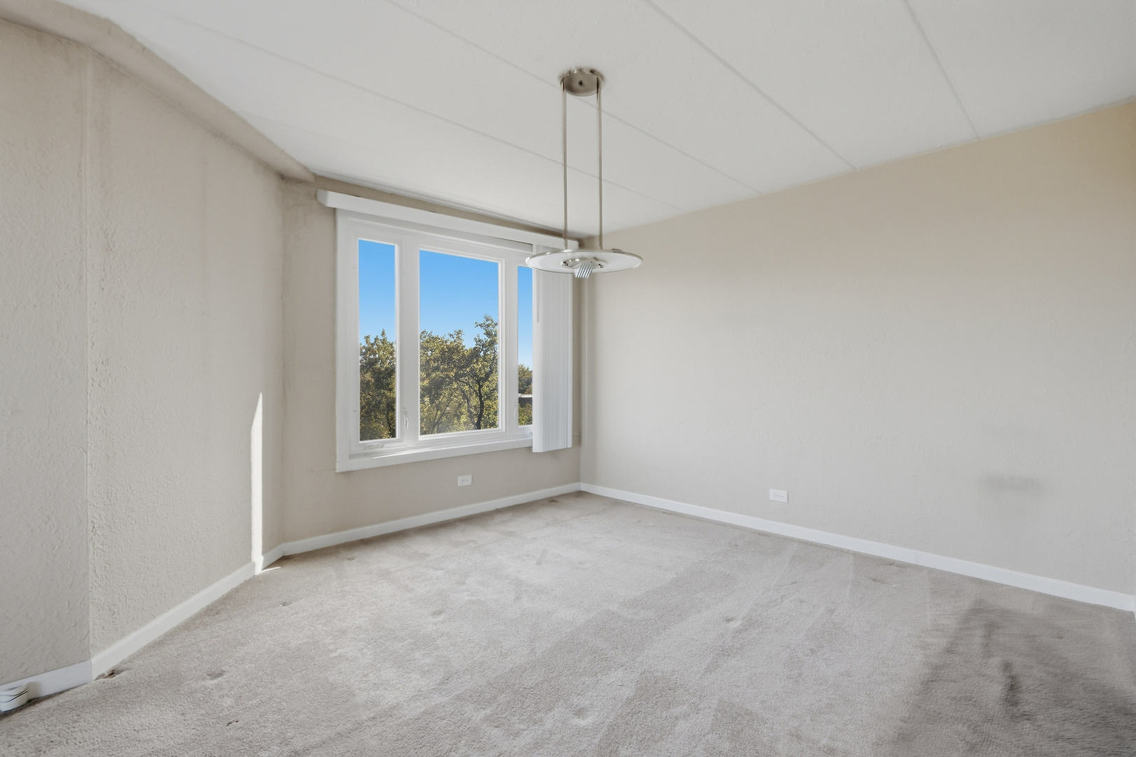 Vacant carpeted room with a large window, vertical blinds, and a hanging ceiling light.