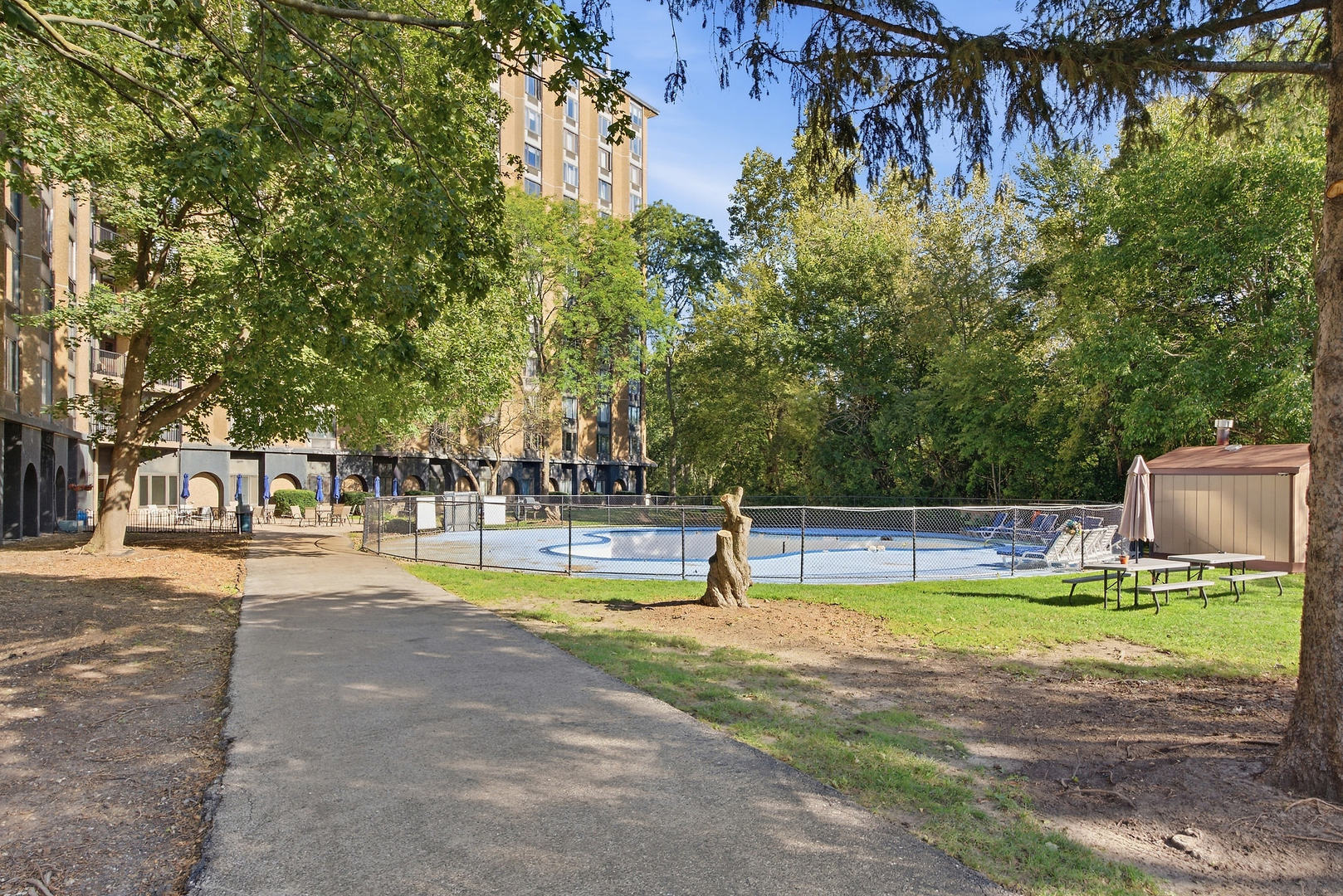 Outdoor pool beside a multi-story apartment building, surrounded by mature trees and a paved walkway.