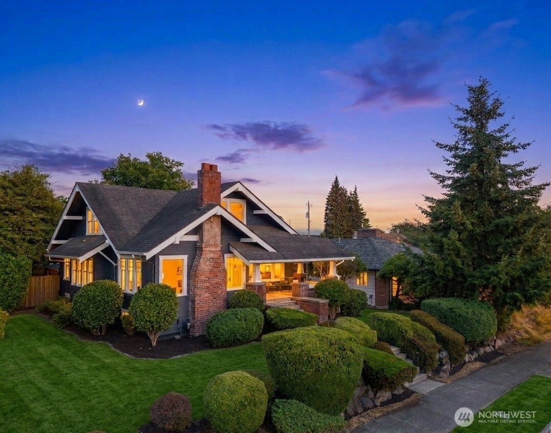 Gabled gray house with a brick chimney, glowing windows, and sculpted front-yard shrubs at dusk.