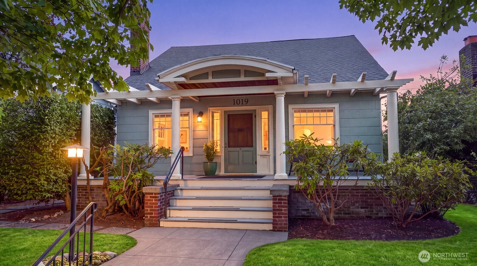 Blue-gray house with a columned porch, broad steps, and warmly lit windows at dusk.