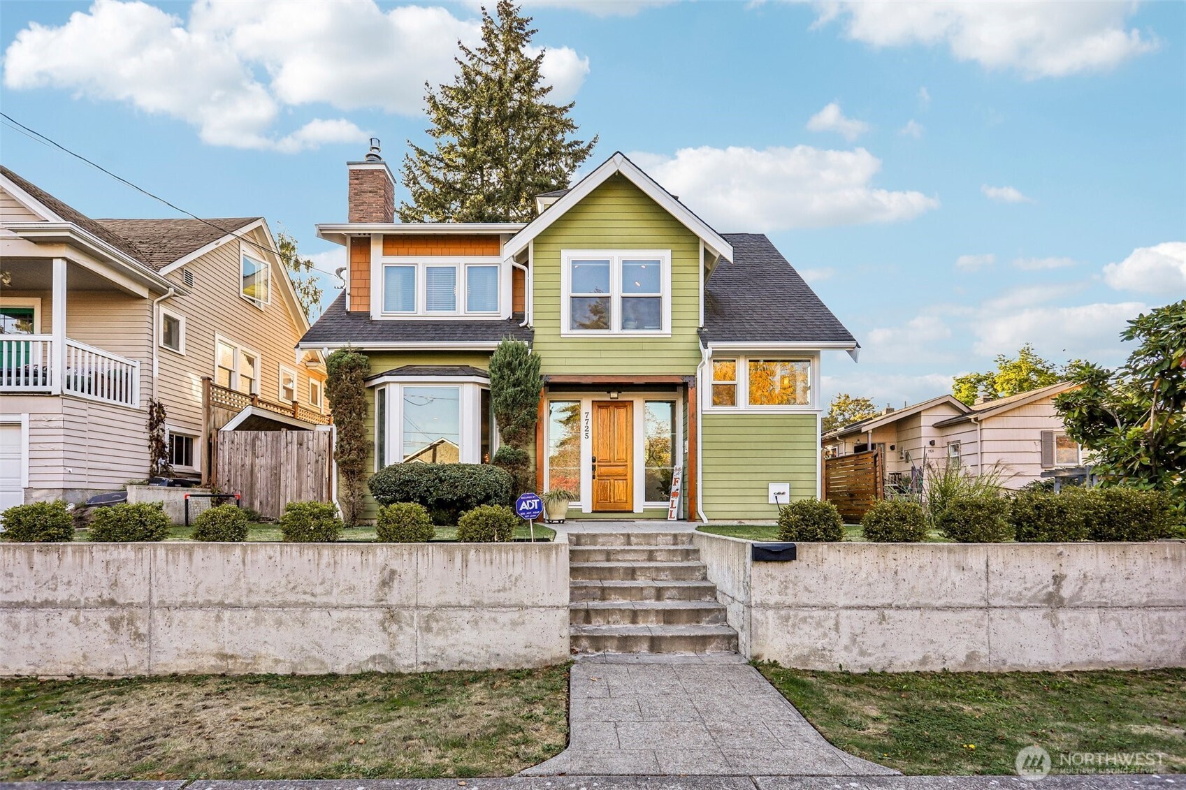 Two-story green house with white trim, a wooden front door, and concrete steps through a retaining wall.