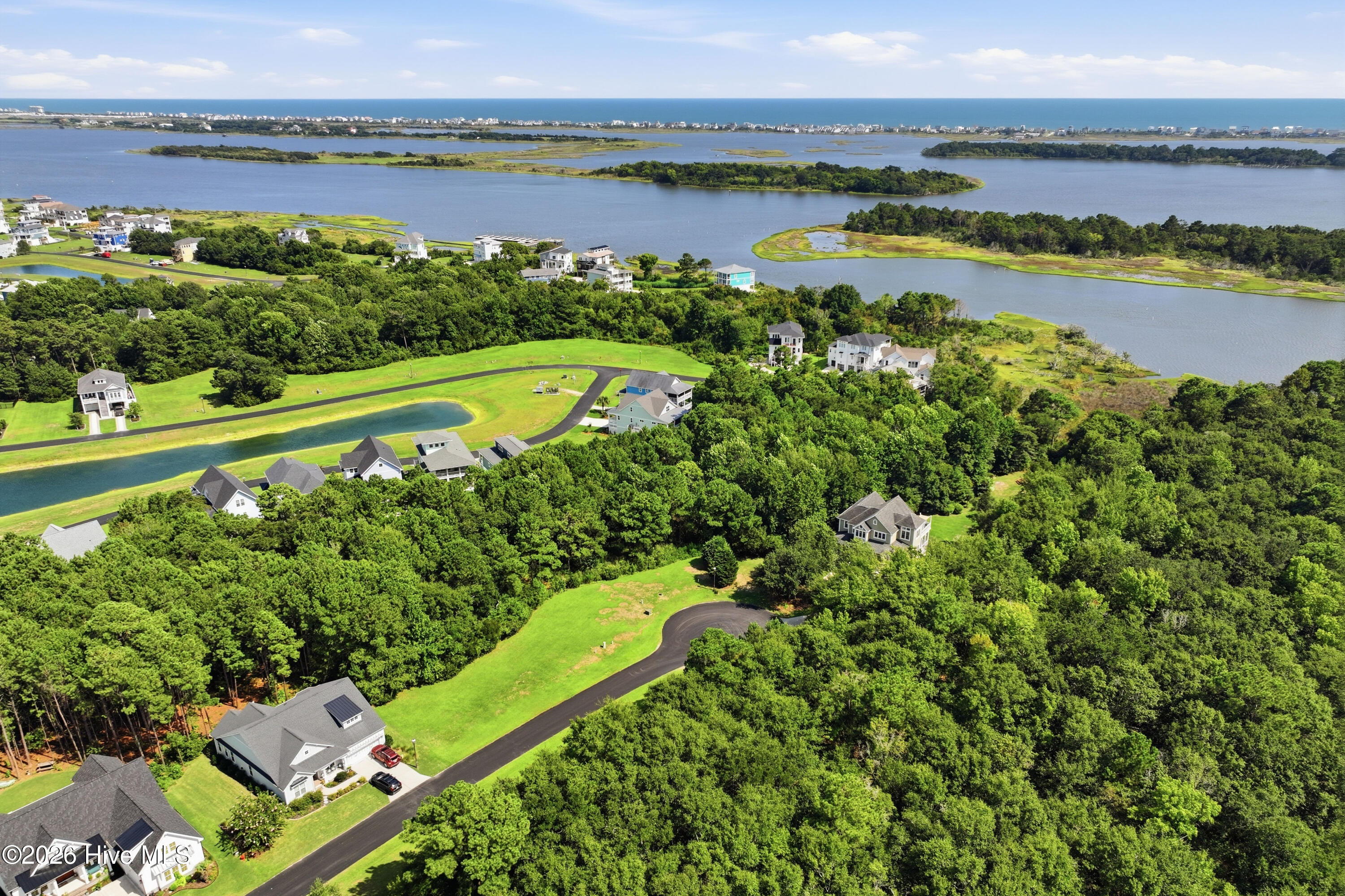 Aerial view of a wooded coastal neighborhood with homes, waterways, marsh islands, and ocean beyond.