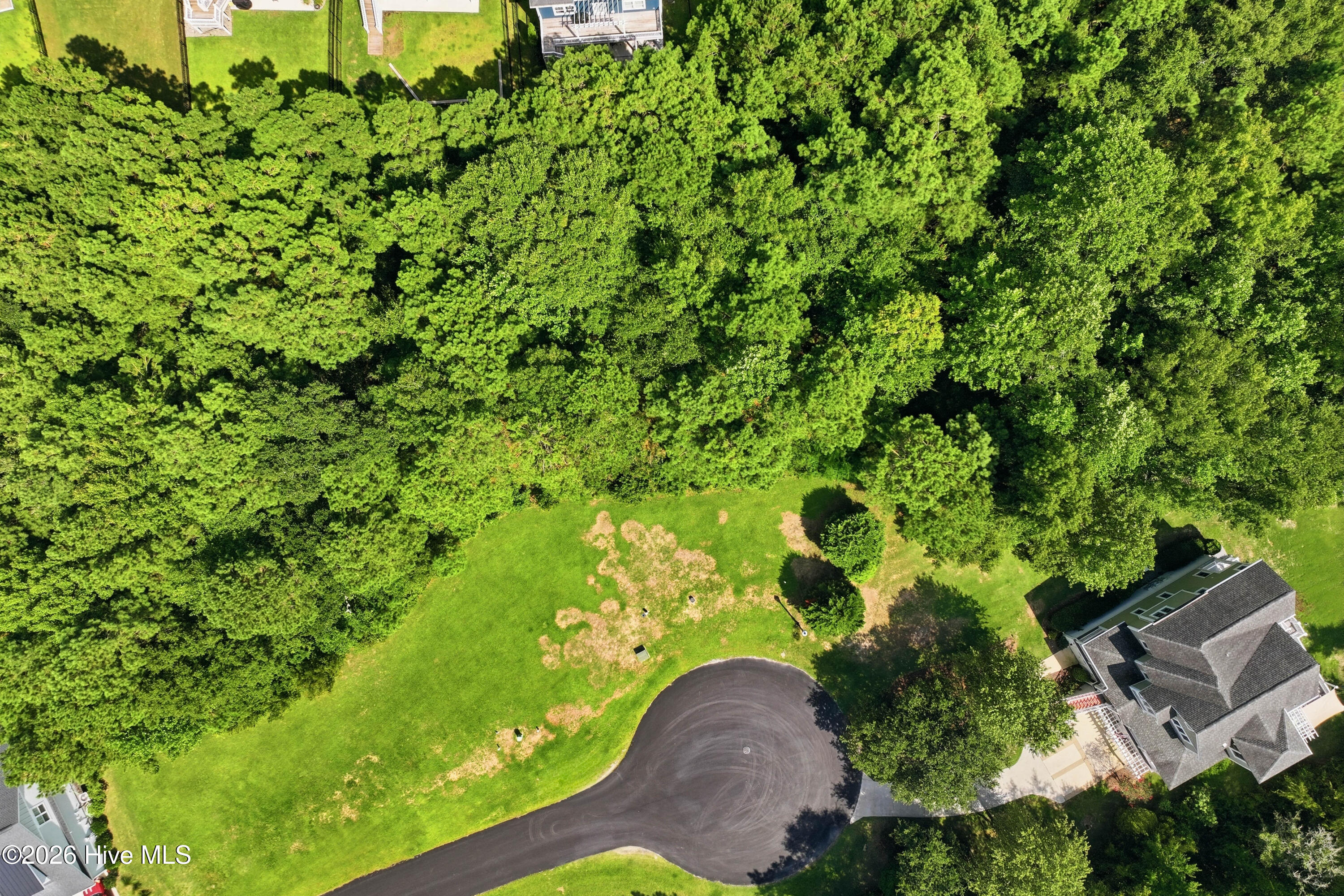 Aerial view of a home beside a large lawn and dense woodland, near a curved cul-de-sac.