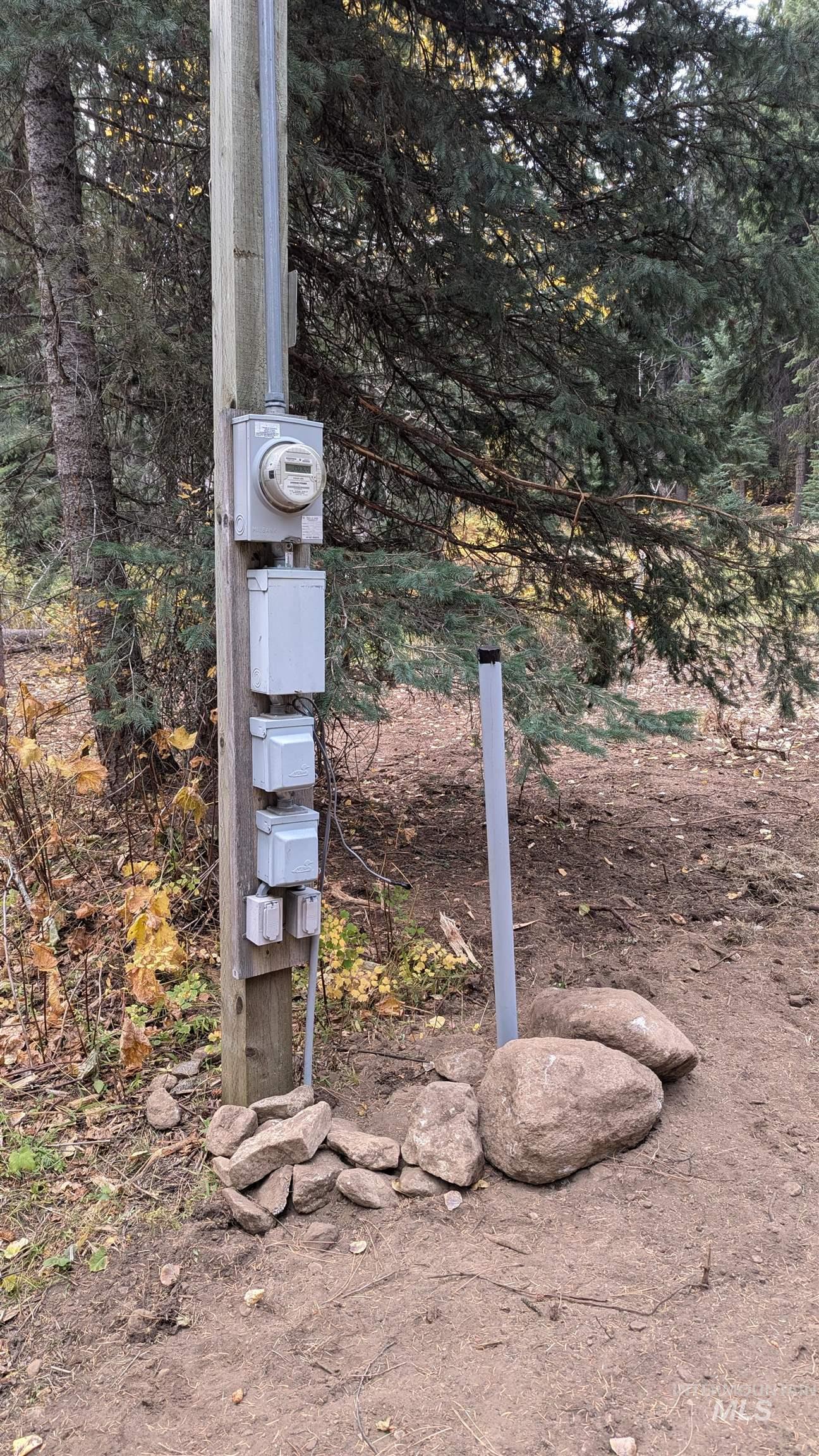 Electrical meter and utility boxes on a pole in a wooded clearing, surrounded by rocks and evergreen trees.