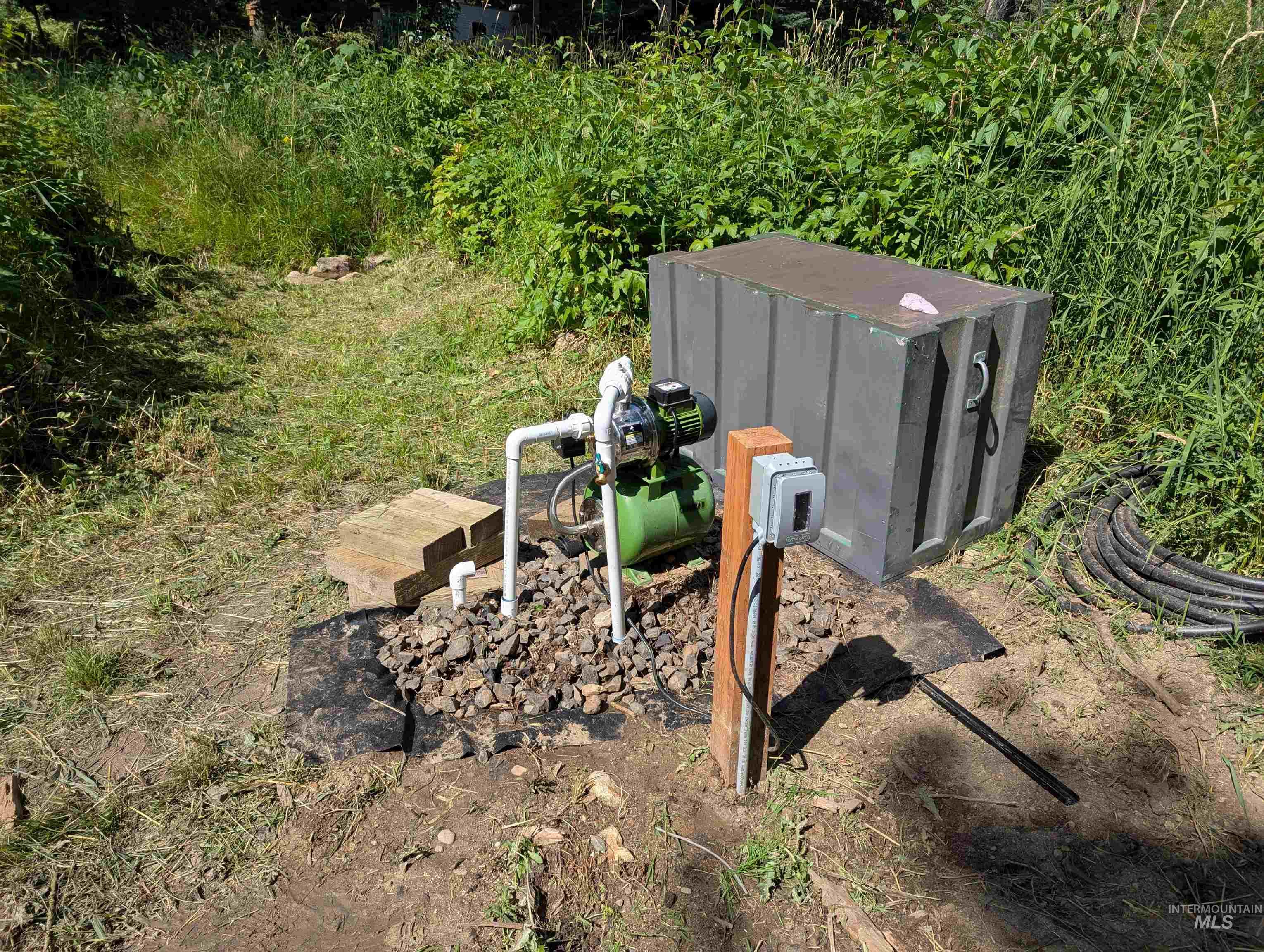 Outdoor water well pump and electrical controls beside a gray utility enclosure.