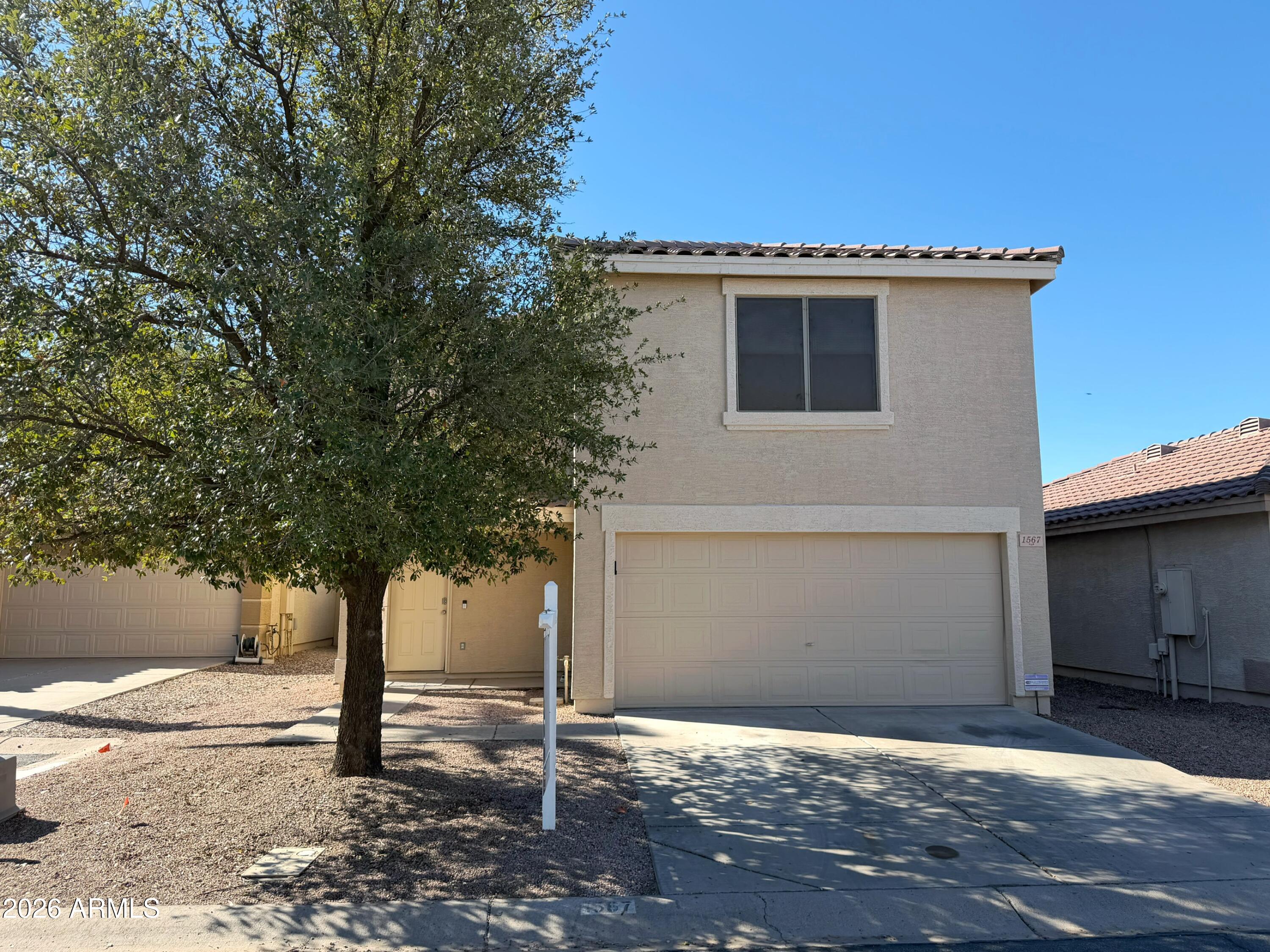 Two-story stucco house with an attached double garage, tile roof, and a large shade tree in front.