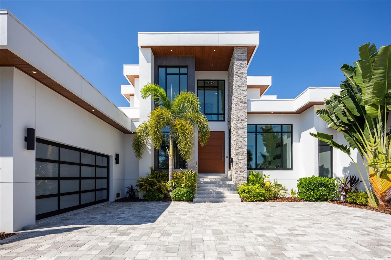 Modern white house with a stone column, tropical landscaping, and a broad paver driveway.