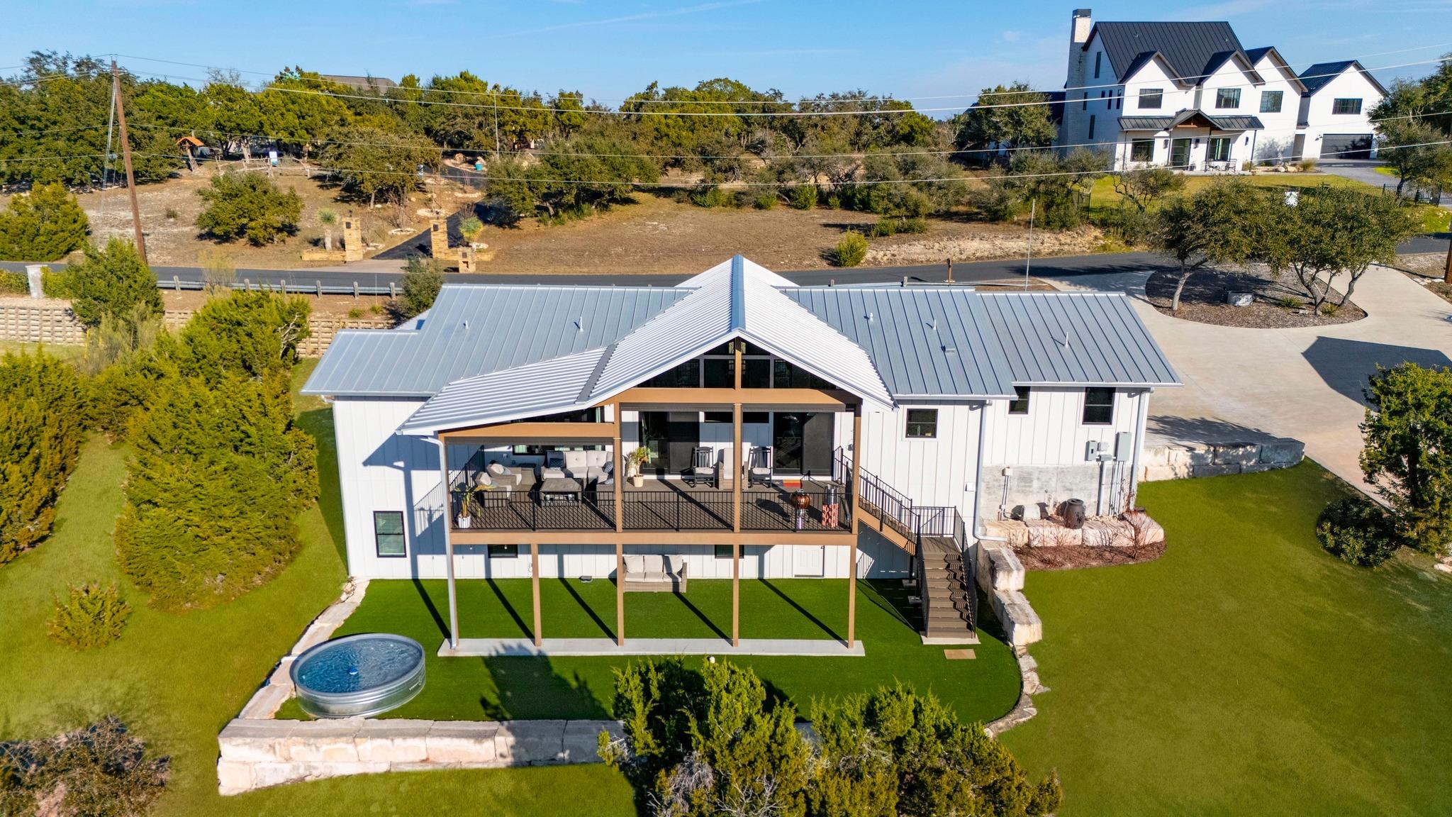 Aerial view of a white home with a raised covered deck, gray metal roof, and round lawn pool.