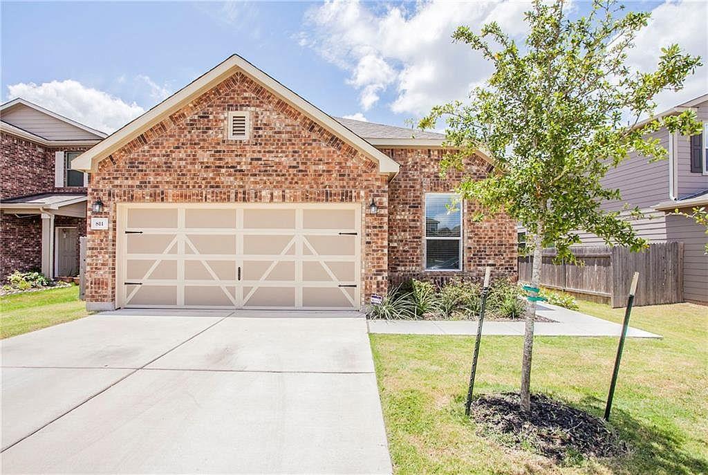 Brick house with a front-gabled roof, double garage, wide driveway, and young tree in the lawn.