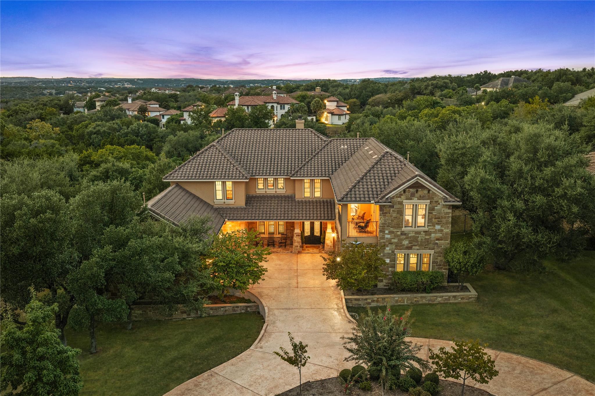 Two-story stone-and-stucco home with a tile roof, glowing windows, and a curved driveway at dusk.