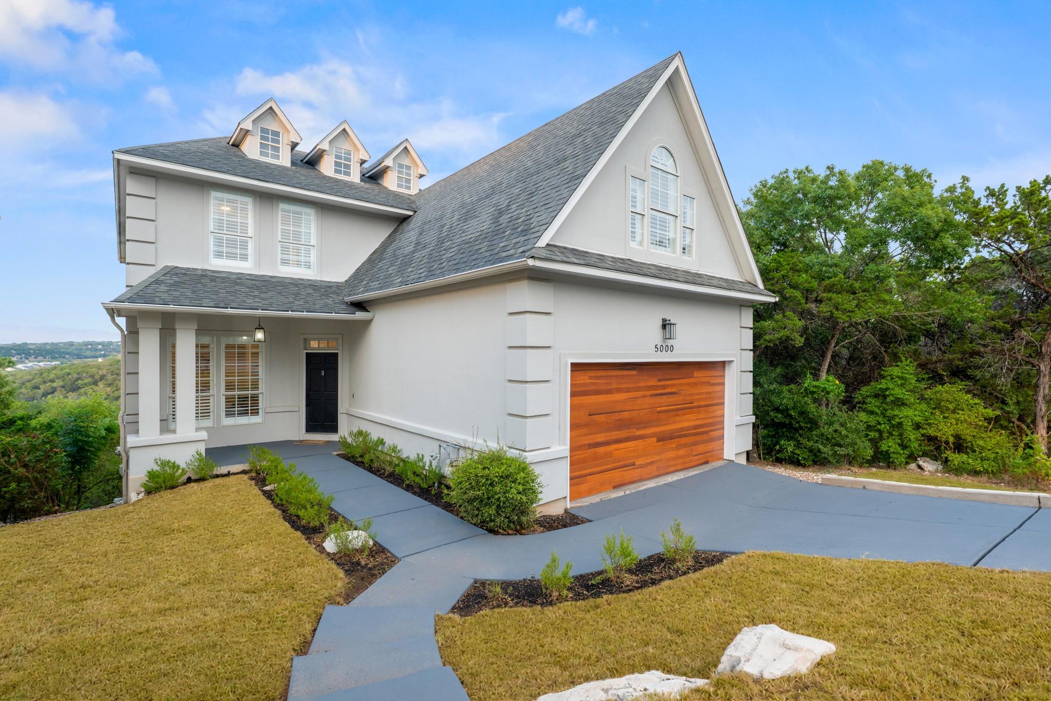 White two-story home with steep gabled roofs, a wood-paneled garage door, and a curved walkway.