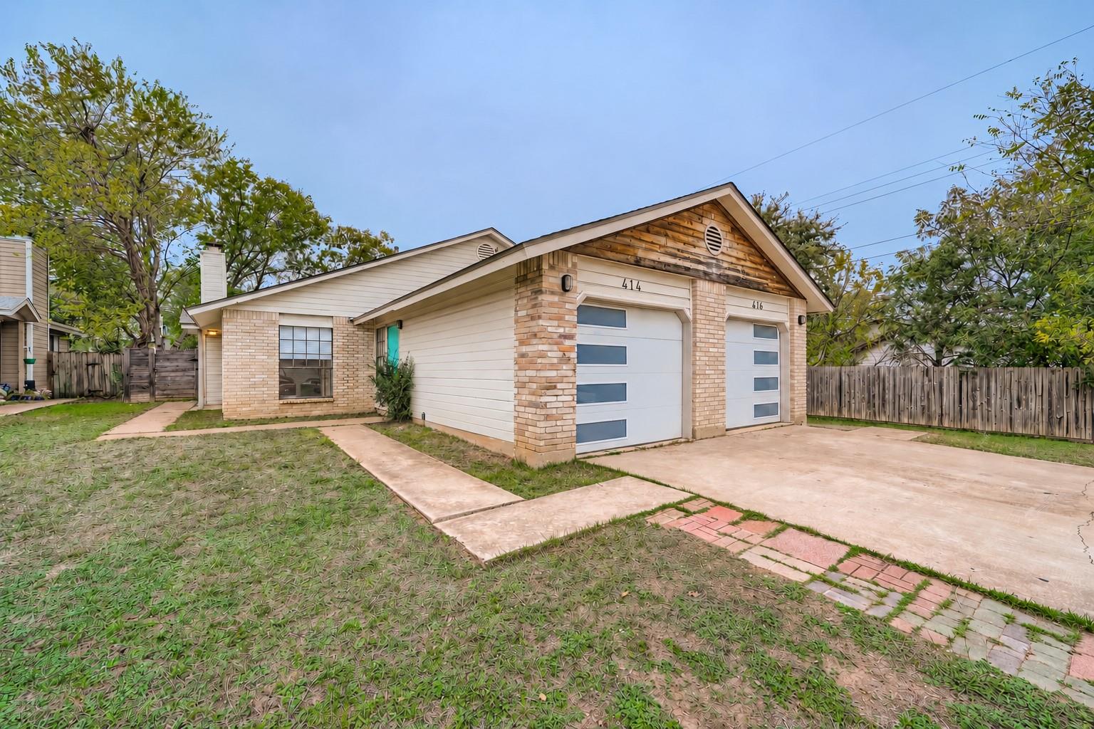 Brick-and-siding building with two white garage doors, a concrete driveway, and fenced front yard.