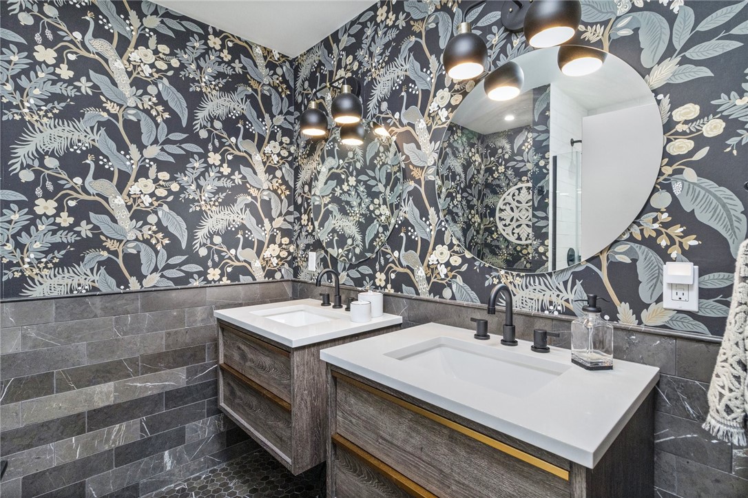 Bathroom with double wood-front vanities, dark floral wallpaper, oval mirrors, and pendant lights.