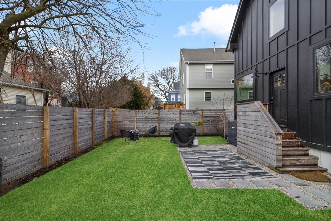 Fenced backyard with a lawn, paved patio, outdoor rug, and steps beside a dark-sided house.