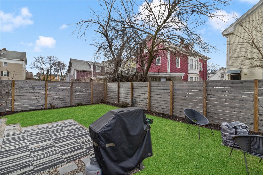 Fenced backyard with a green lawn, striped patio rug, covered grill, and outdoor seating.