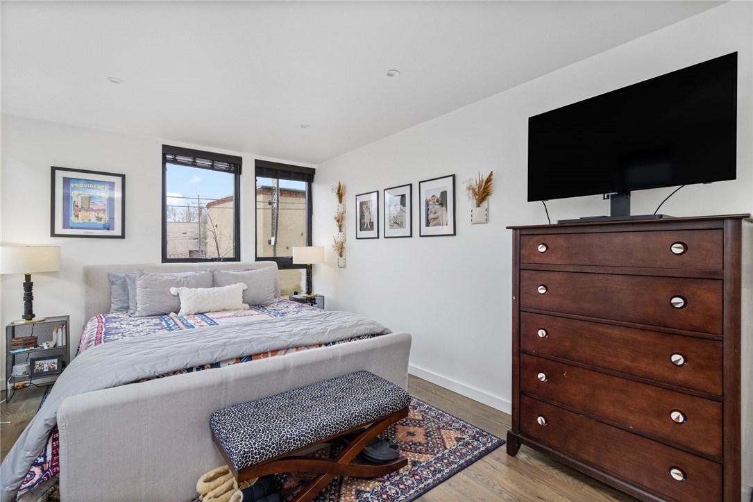 Furnished bedroom with a gray upholstered bed, patterned rugs, tall windows, and a dark wood dresser.