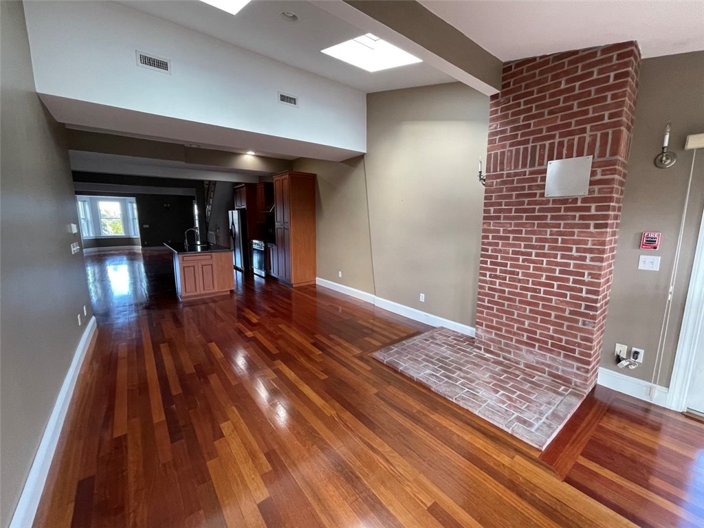 Open living area with hardwood floors, a brick fireplace, skylights, and the kitchen beyond.