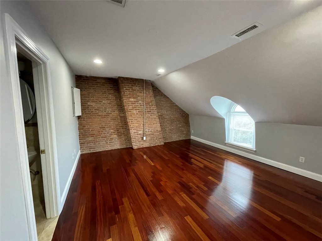 Empty attic room with polished hardwood floors, exposed brick, sloped ceiling, and an arched dormer window.