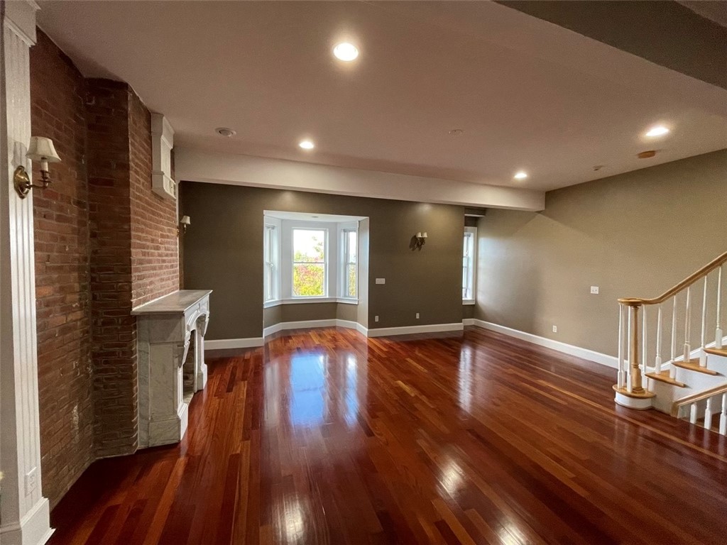 Empty living room with glossy hardwood floors, exposed brick fireplace, bay window, and staircase.