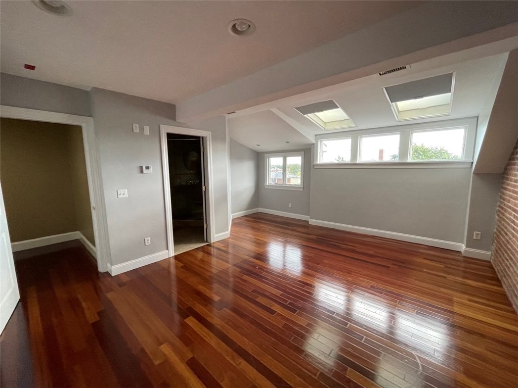 Empty living room with glossy hardwood floors, skylights, broad windows, and an exposed brick wall.