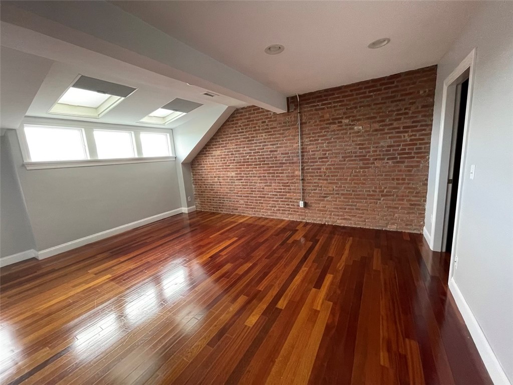 Empty loft-style room with polished hardwood floors, exposed brick wall, skylights, and wide windows.