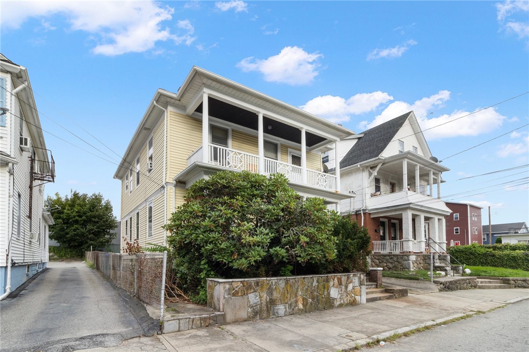 Yellow two-story house with a covered upper porch, front shrubs, and a stone retaining wall.