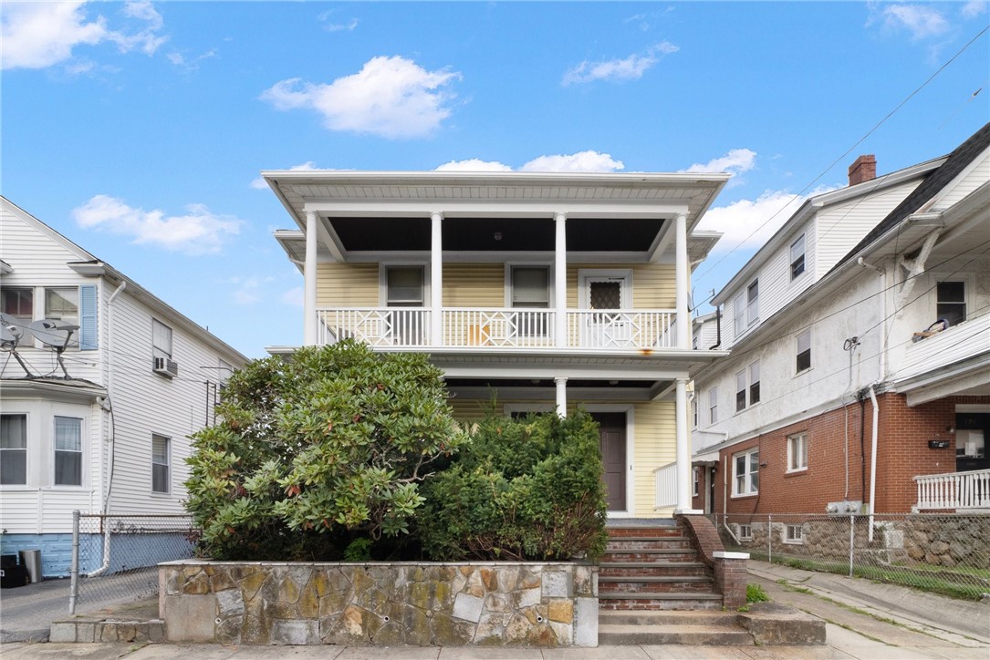 Yellow two-story house with a covered porch, upper balcony, and stone wall along the front yard.
