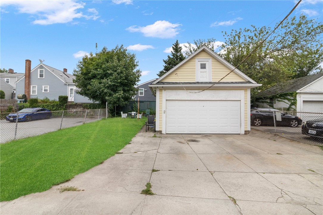 Detached pale-yellow garage with a white double door, broad concrete driveway, and fenced lawn.