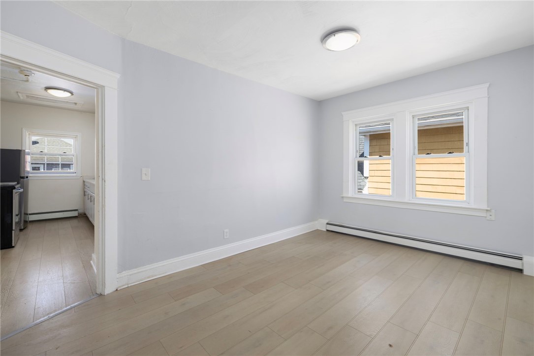 Empty light-gray room with pale wood floors, two windows, and an open doorway to the kitchen.