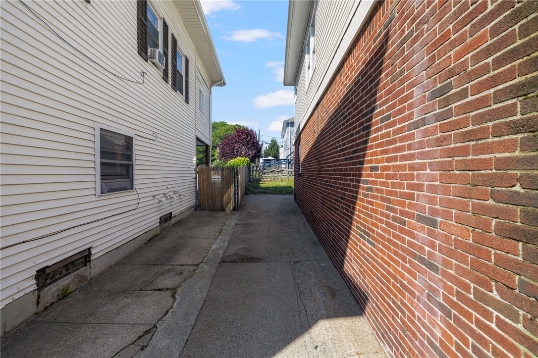 Paved passage between a white-sided house and brick building, leading to a fenced yard.