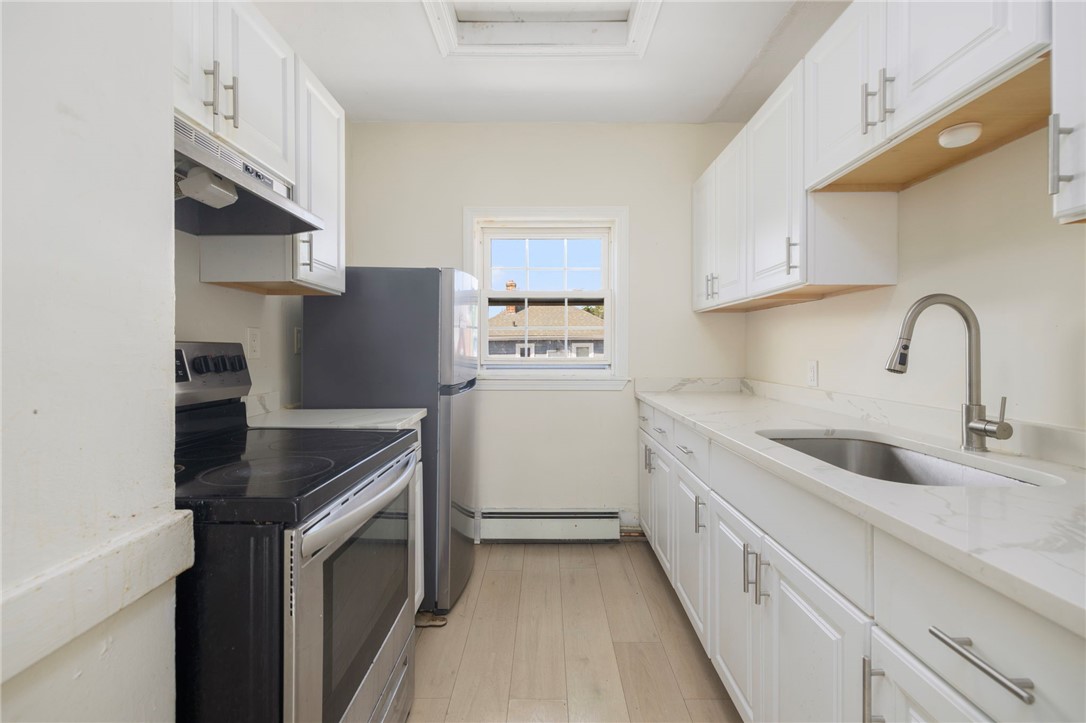 Narrow kitchen with white cabinets, a black range, stainless-steel refrigerator, and sink beneath a window.