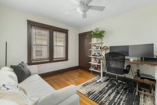 Home office with a desk and two monitors beside a white sofa and large window.