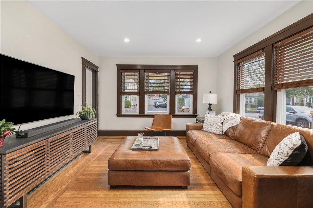 Living room with a brown leather sectional, matching ottoman, hardwood floors, and large windows.