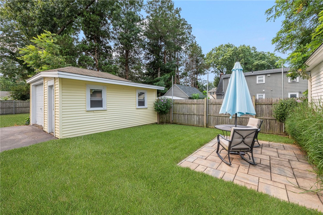 Fenced backyard with a yellow shed and stone patio furnished with chairs and a blue umbrella.