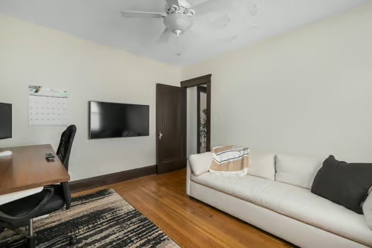 Living room with a white sofa, wall-mounted TV, desk, and hardwood floors.