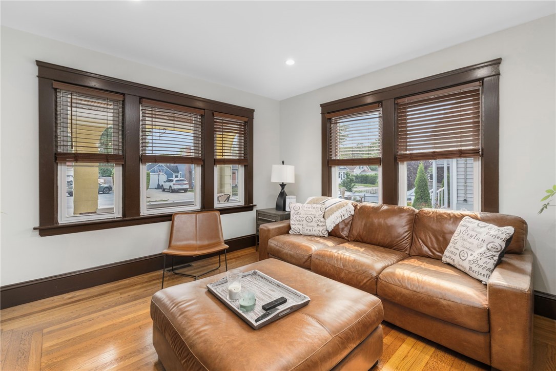 Living room with a brown leather sofa, matching ottoman, hardwood floors, and large windows with wooden blinds.