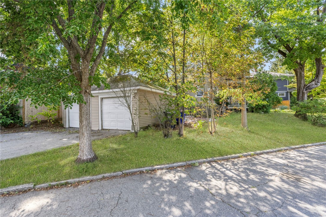 Detached beige garage and driveway beneath mature trees beside a leafy lawn.
