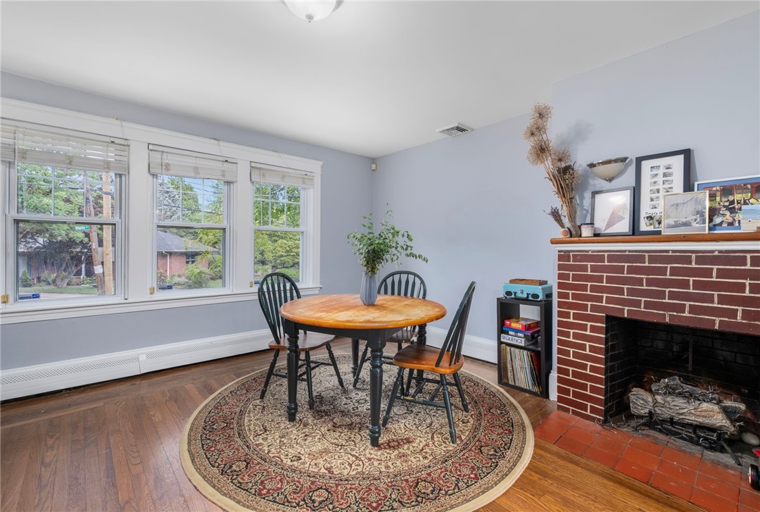 Dining room with a round wooden table, patterned rug, three large windows, and a brick fireplace.