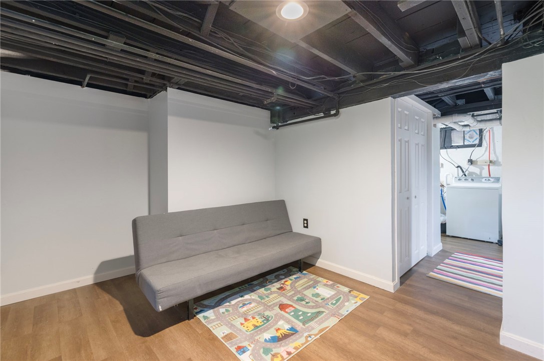 Basement living area with a gray futon, colorful play rug, exposed ceiling, and adjoining laundry area.