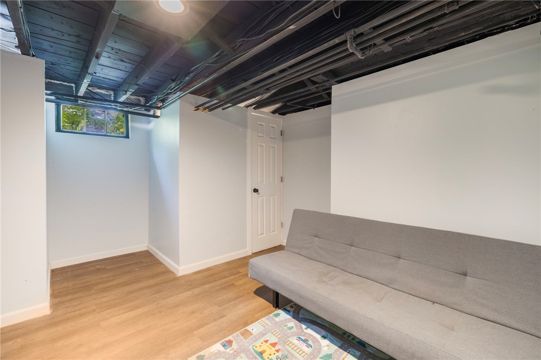 Finished basement room with a gray sofa, colorful rug, exposed ceiling joists, and a small window.