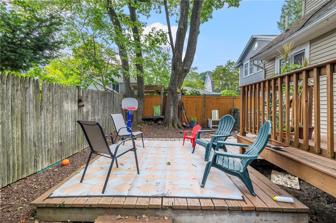 Fenced backyard patio with outdoor seating, a wooden deck, mature trees, and a basketball hoop.