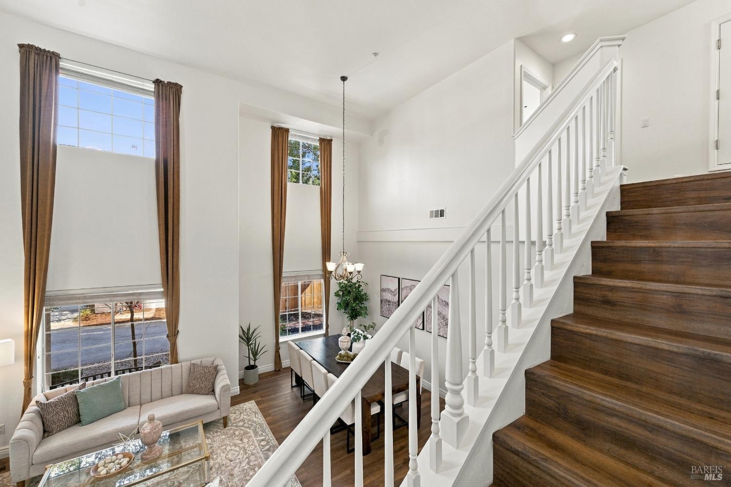 Furnished two-story living room with tall windows and a dark wood staircase with white railings.