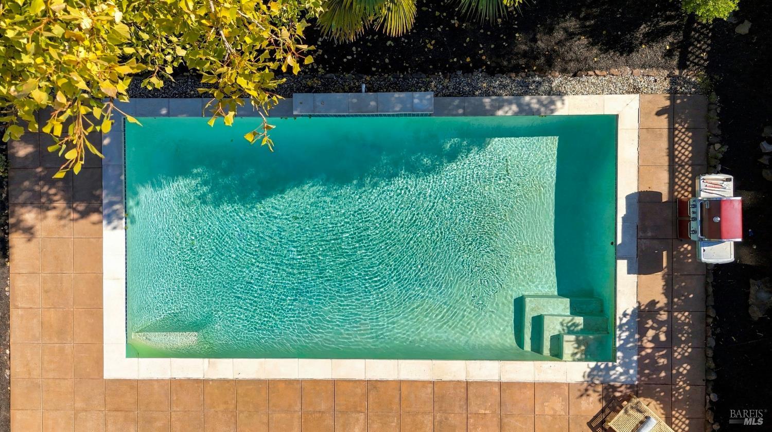 Swimming pool with built-in steps, tan tiled patio, and a grill, viewed from above.