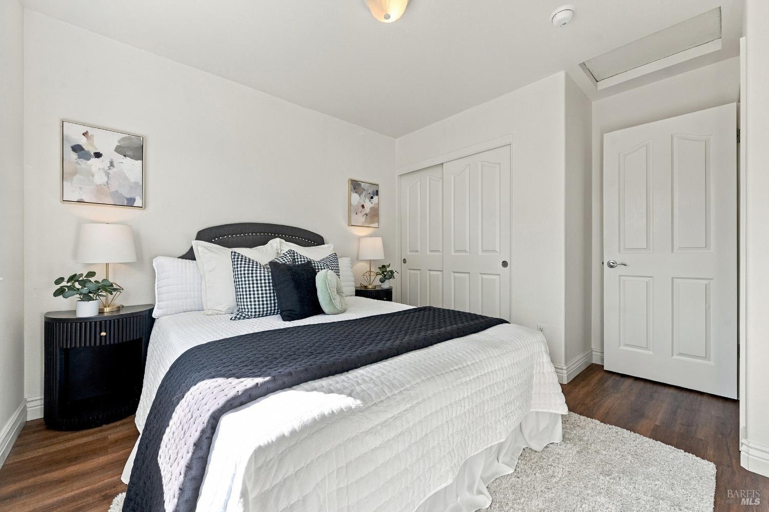 Staged bedroom with white bedding, a charcoal throw, dark nightstands, and a white shag rug.