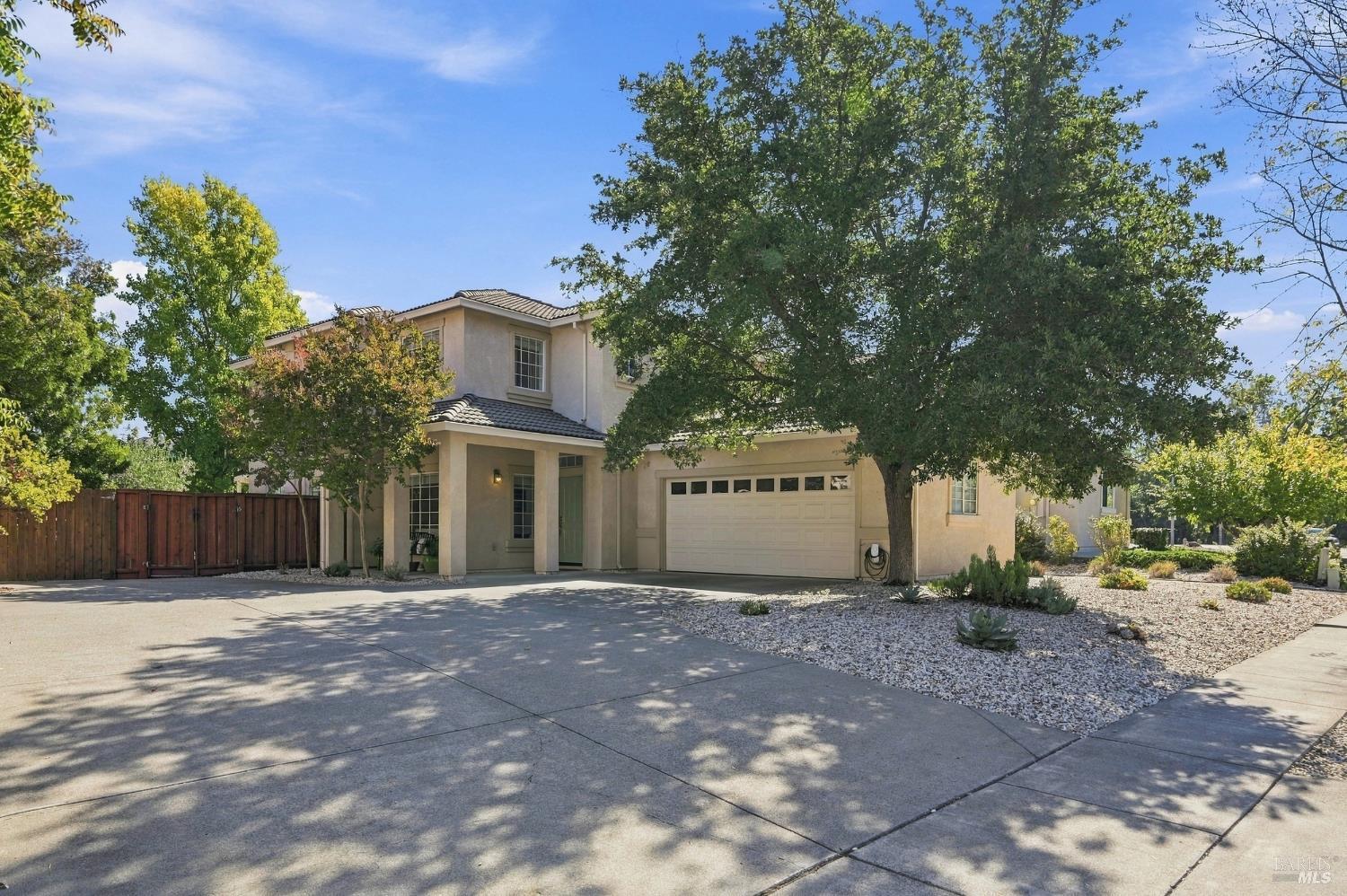 Two-story beige stucco home with a tiled roof, attached garage, and broad driveway beneath mature trees.