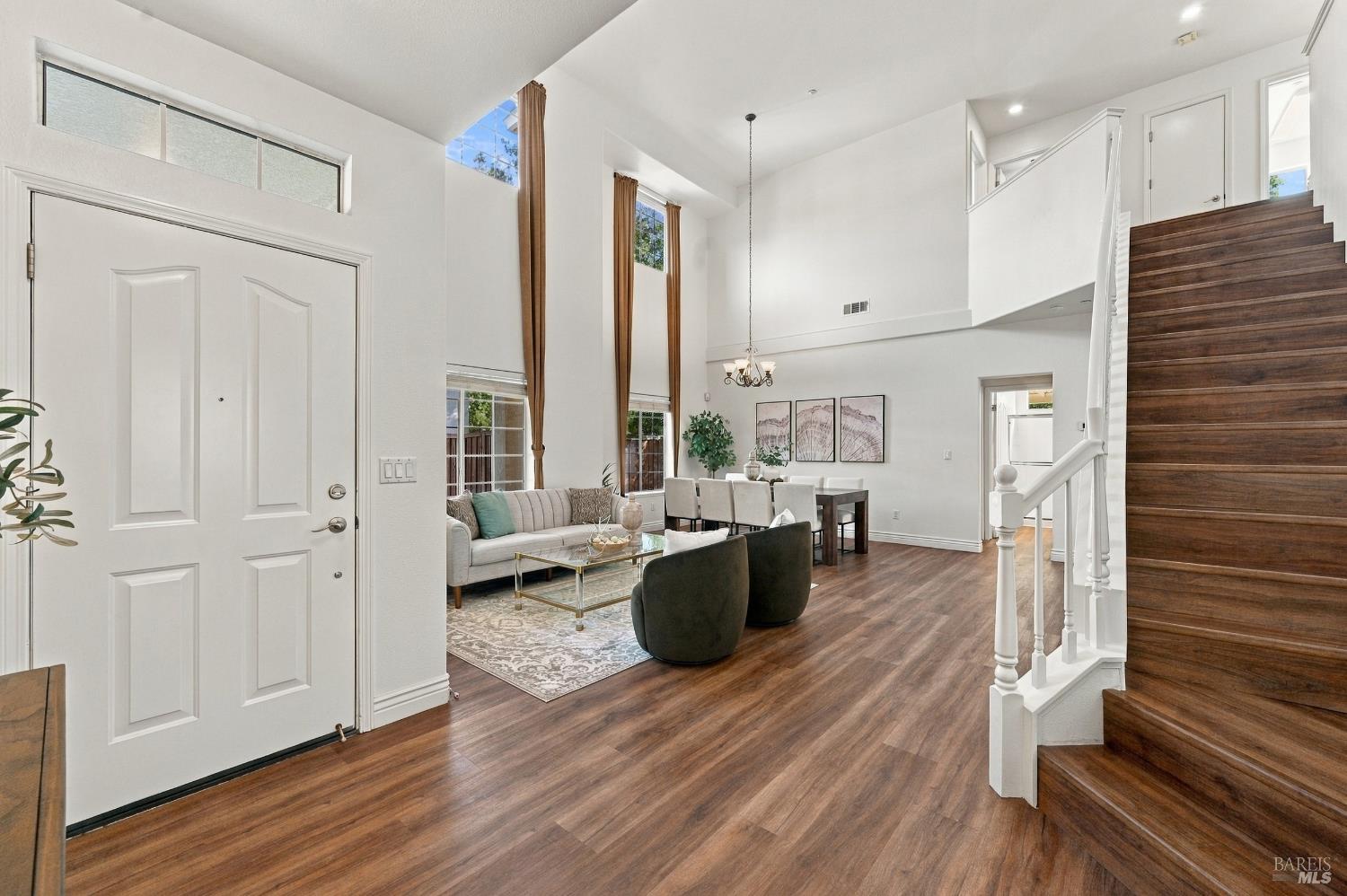 Open living and dining room with a vaulted ceiling, tall windows, and a dark wood staircase.