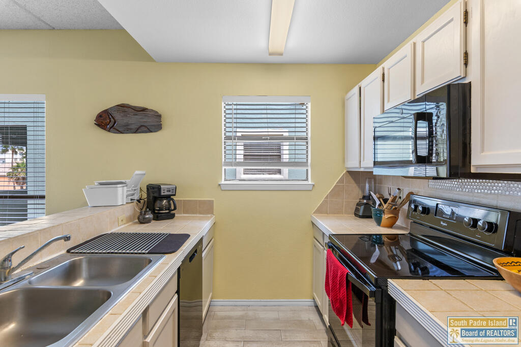 Galley kitchen with white cabinets, tiled counters, stainless steel sink, and black appliances.